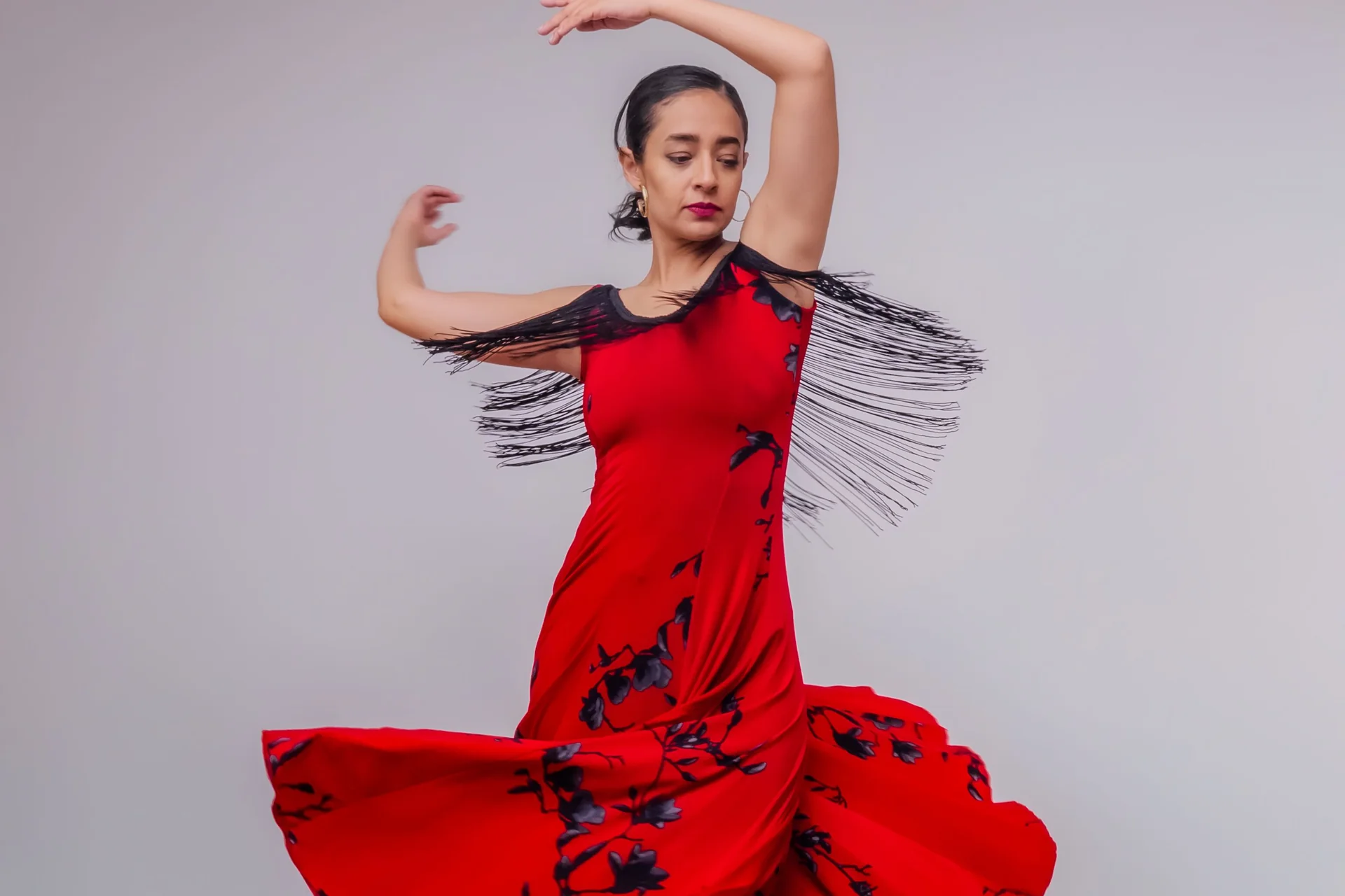 A flamenco dancer in a red floral-print dress with fringed shoulder straps, arms raised mid-turn against a plain grey backdrop